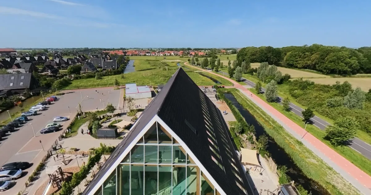 Een schoolgebouw met een glazen puntgevel aan een sloot en een fietspad, gezien vanuit de drone